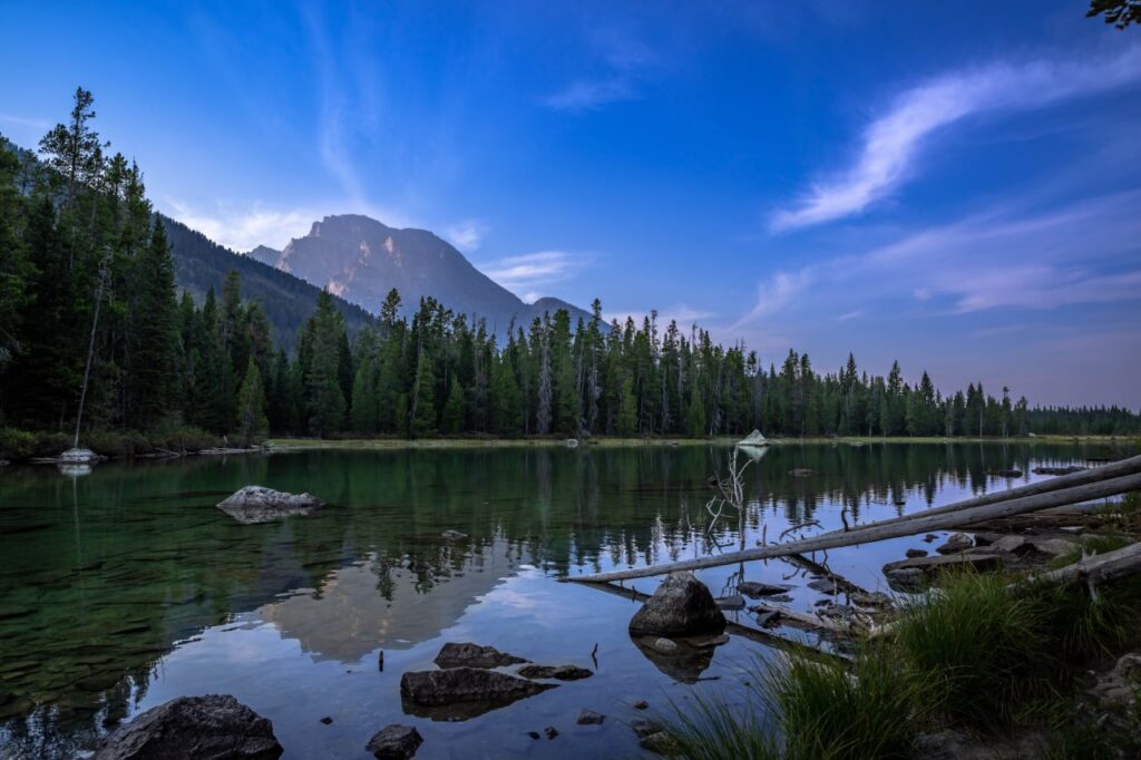 Quiet mountain lake reflecting the Teton Range and surrounding evergreen forest beneath a blue evening sky in Grand Teton National Park. Copy
